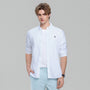 A male model wearing a white long-sleeve button-down shirt with a small embroidered logo on the chest, layered over a white t-shirt and paired with light blue pants, against a solid gray background.